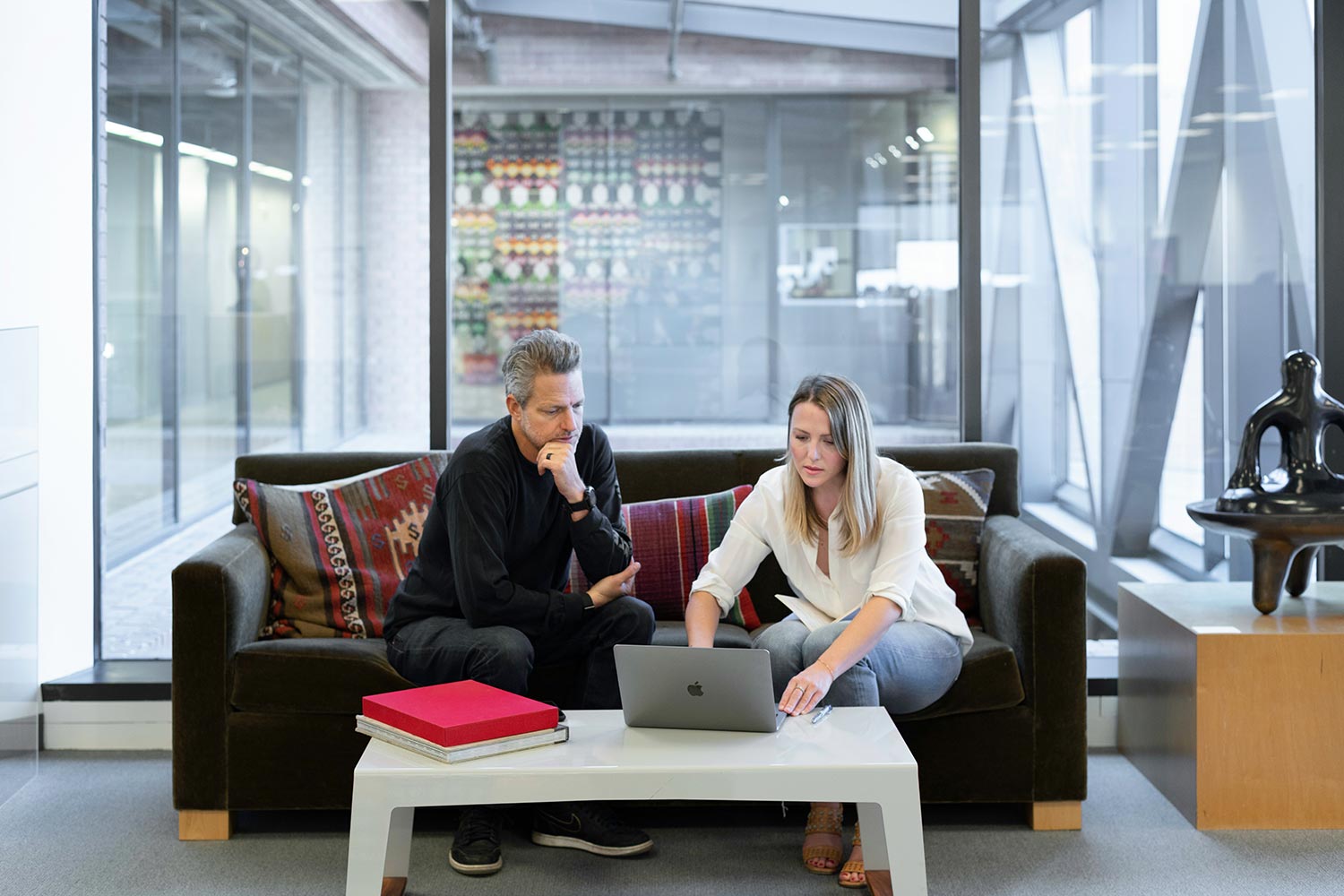 Two people sitting on a couch collaborating over a laptop in a modern office