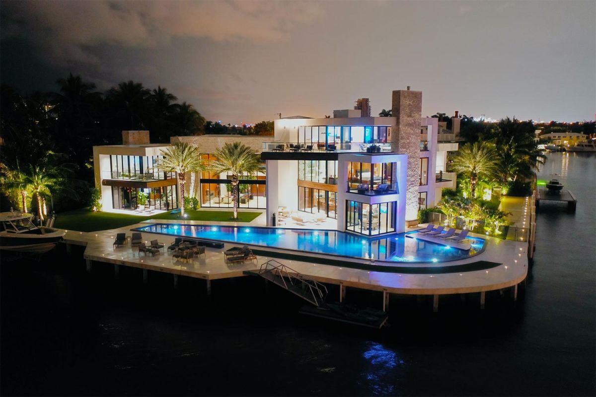 Luxury waterfront mansion illuminated at night with pool and palm trees